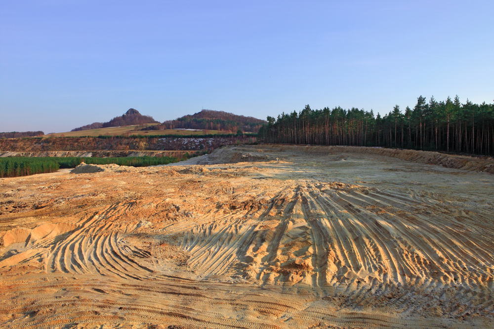 A large, sandy clearing with visible vehicle tire tracks, bordered by a forest and distant hills under a clear sky.