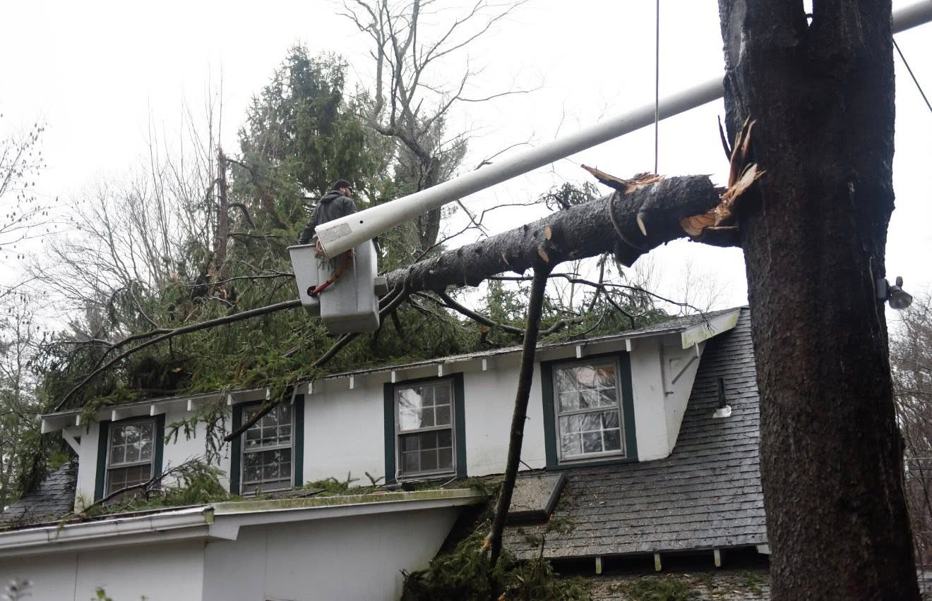 A worker in a bucket lift removes a large tree that has fallen onto the roof of a house, breaking utility lines and causing visible damage.