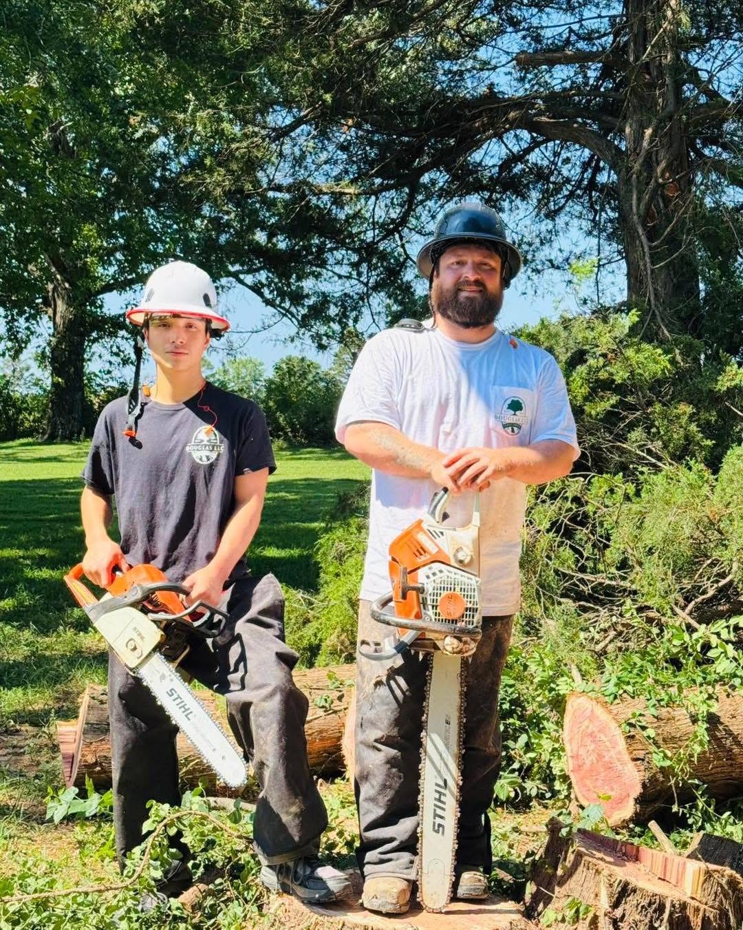 Two people wearing protective helmets and work clothes stand outdoors holding chainsaws, surrounded by cut tree branches and logs.